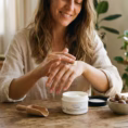 Mujer sonriente aplicando crema de manos Koken en su hogar, con tarro 250ml sobre mesa de madera y nueces de karité en ambiente natural y acogedor